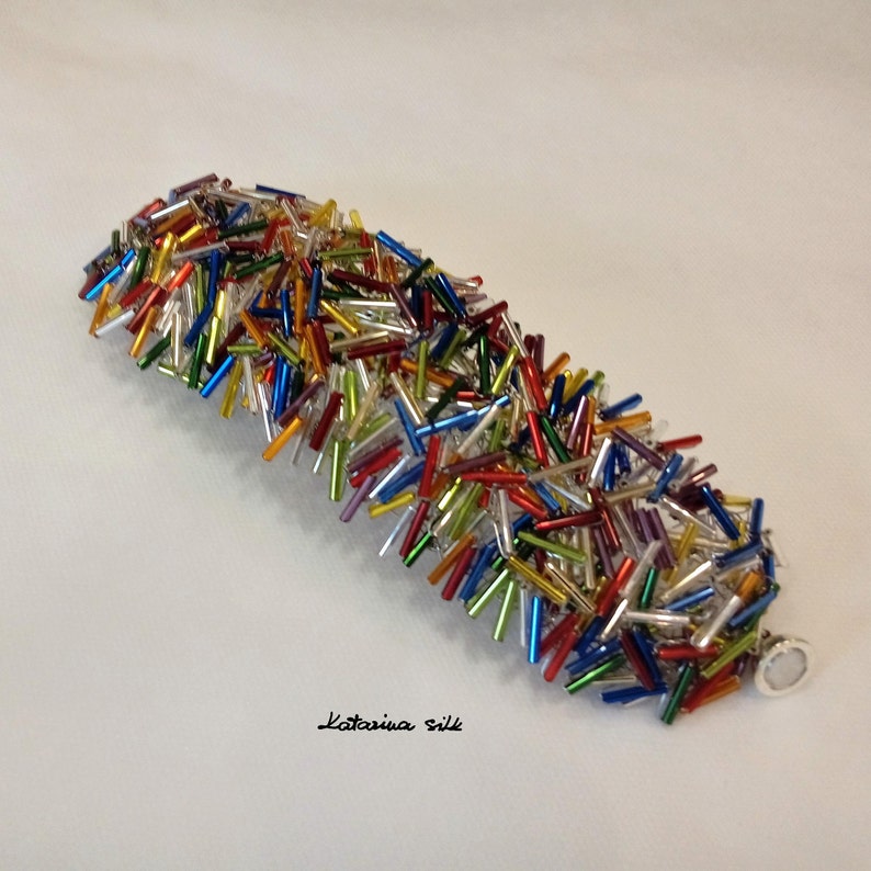 May include: A colorful bracelet made of small glass beads in various colors, including red, blue, green, yellow, and silver. The bracelet is made of a single strand of beads and has a silver clasp.