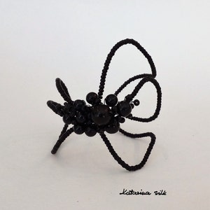 May include: Black beaded spider with long legs made of wire. The spider is posed on a white surface.