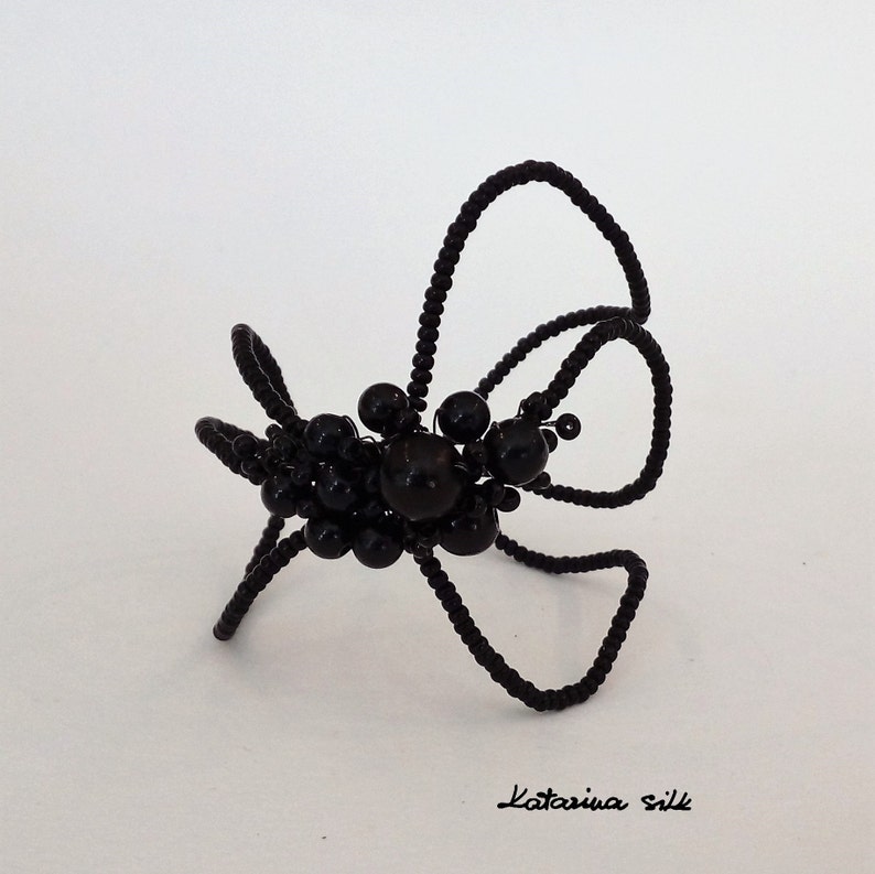 May include: Black beaded spider with long legs made of wire. The spider is posed on a white surface.