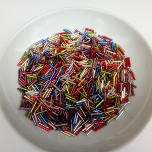 May include: A bowl filled with colorful glass beads. The beads are small and cylindrical, with a metallic sheen. The colors include red, gold, green, blue, and purple.