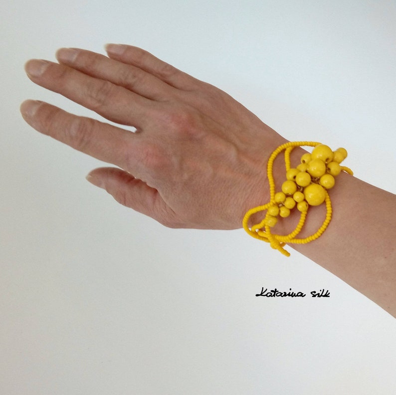 May include: A bright yellow beaded bracelet with a cluster of larger beads in the center. The bracelet is worn on a person's wrist.