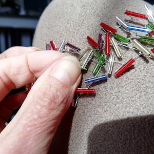 May include: A close-up of a hand attaching colorful glass beads to a brown felt hat. The beads are arranged in a pattern and are connected by silver wire.
