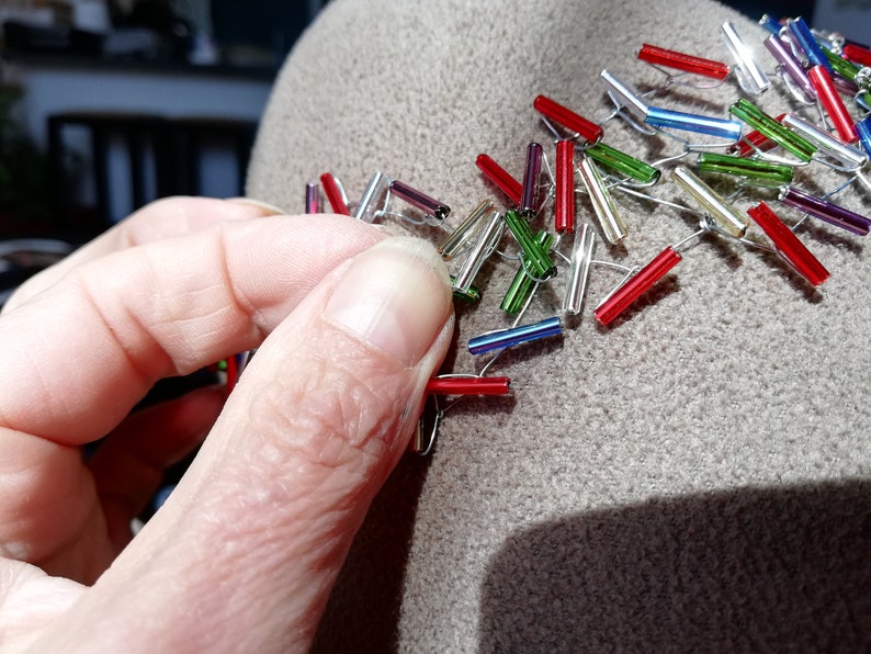 May include: A close-up of a hand attaching colorful glass beads to a brown felt hat. The beads are arranged in a pattern and are connected by silver wire.