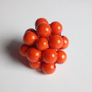 May include: A cluster of orange wooden beads are wired together to form a sphere. The beads are all the same size and are arranged in a random pattern.