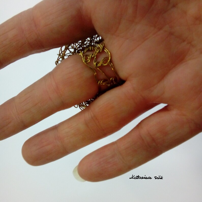 May include: A gold wire ring with a delicate, intricate design. The ring is worn on a finger and the back of the hand is visible.