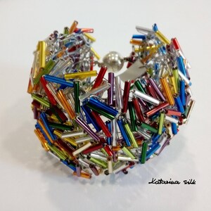 May include: A colorful bracelet made with small glass tubes and silver wire. The bracelet is a mix of red, orange, yellow, green, blue, purple, and silver.
