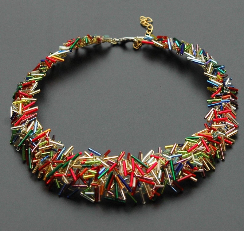 May include: A colorful necklace made of small, rectangular beads in shades of red, green, blue, gold, and silver. The beads are arranged in a random pattern, creating a unique and eye-catching design.
