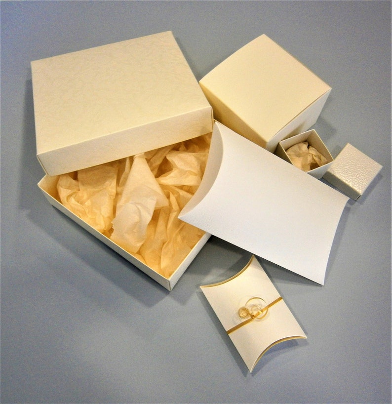 May include: A set of five white gift boxes, including a large square box with a lid, a smaller square box with a lid, a pillow box with a lid, and two smaller square boxes without lids. The boxes are filled with white tissue paper.