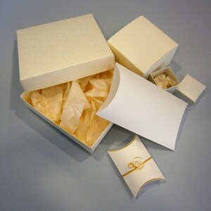 May include: A set of five ivory-coloured gift boxes, including a larger box with a lid and tissue paper inside, a smaller box with a lid, and two pillow boxes. The pillow boxes are open and show a gold ribbon inside.