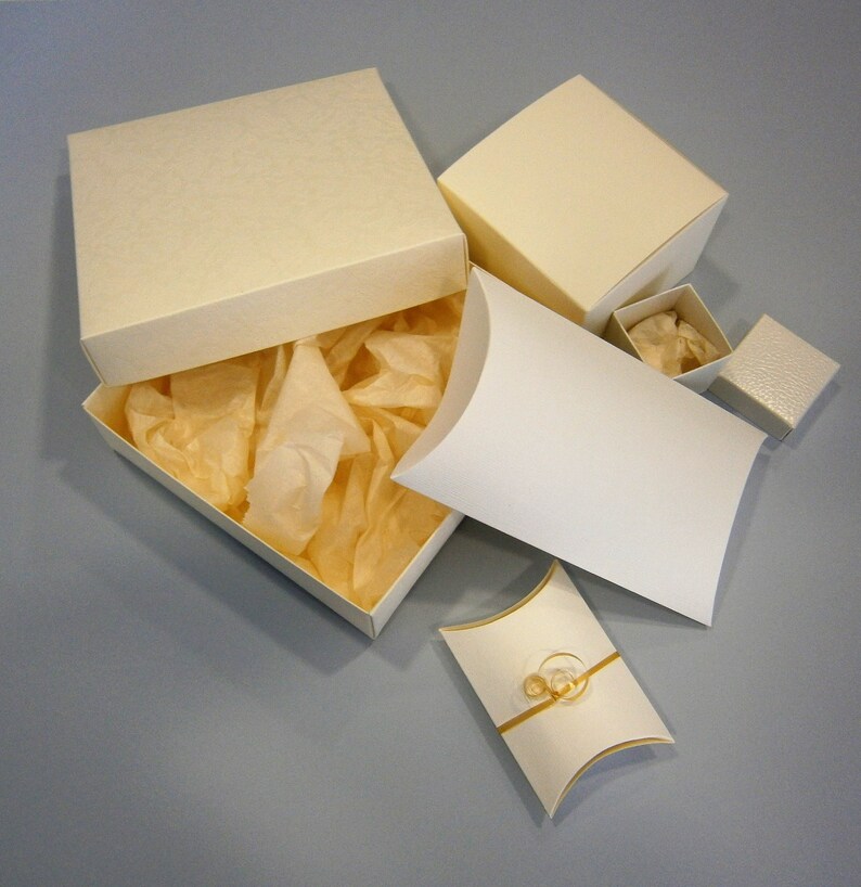 May include: A set of five ivory-coloured gift boxes, including a larger box with a lid and tissue paper inside, a smaller box with a lid, and two pillow boxes. The pillow boxes are open and show a gold ribbon inside.