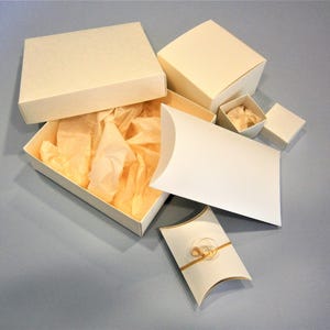 May include: A set of five white gift boxes, including a square box with a lid, a rectangular box with a lid, a pillow box, and two smaller square boxes. The boxes are made of sturdy cardboard and have a smooth, matte finish.