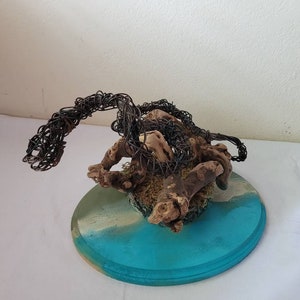 May include: A wire sculpture of a dragon with a brown driftwood base, set on a round wooden base painted with blue and white paint.