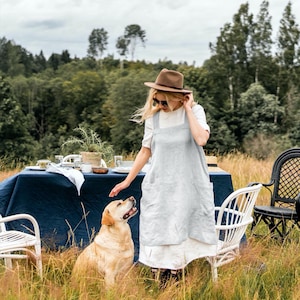 May include: A woman wearing a light blue linen apron and a brown hat stands in a field of tall grass. She is petting a golden retriever dog. A blue tablecloth covers a table with white chairs in the background.