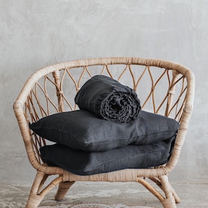 Könnte beinhalten: Eine schwarze Leinen-Decke und zwei passende Kissen liegen auf einem geflochtenen Korbstuhl. Der Stuhl steht auf einem Naturfaser-Teppich mit einem weißen Muster.