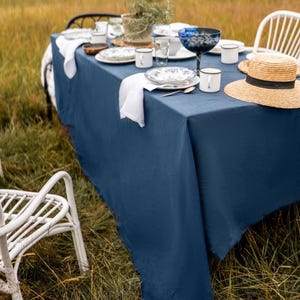 May include: A blue linen tablecloth set on a table in a field with white chairs, plates, cups, and a straw hat.