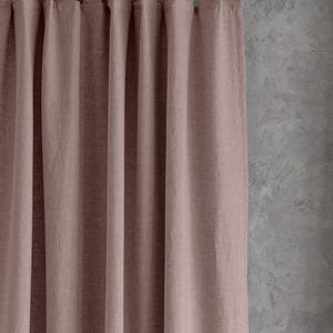Op de afbeelding: Een stoffig roze linnen gordijn met een roede-zak bovenaan en een geplooide look. Het gordijn hangt tegen een getextureerde grijze muur.