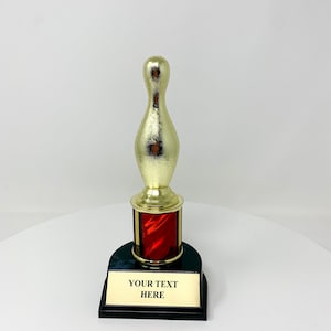 May include: A gold-colored bowling pin trophy with a red and black base. The trophy has a gold-colored pin and a black base with a gold plate that reads "YOUR TEXT HERE".