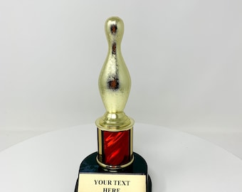 May include: A gold-colored bowling pin trophy with a red and black base. The trophy has a gold-colored pin and a black base with a gold plate that reads "YOUR TEXT HERE".