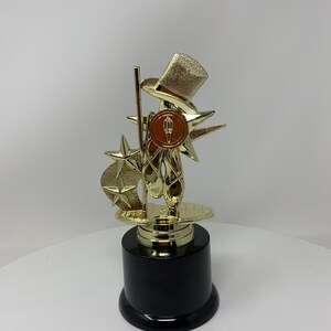 May include: Gold-colored trophy with a black base. The trophy features a top hat, a star, and a pair of ballet shoes.