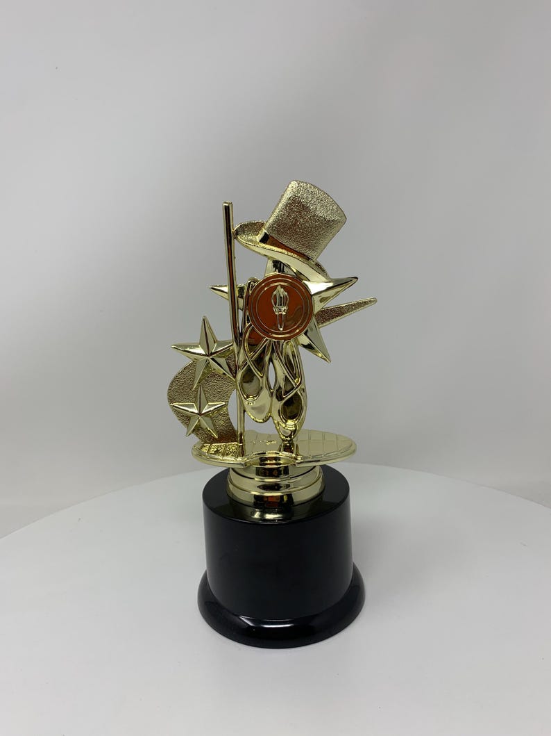 May include: Gold-colored trophy with a black base. The trophy features a top hat, a star, and a pair of ballet shoes.