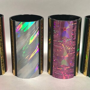 Może przedstawiać: Cztery ozdobne cylindry z iryzującymi wykończeniami. Cylindry są czarne z pozłacanymi obramowaniami i mają r&oacute;żne holograficzne wzory: czerwony, biały, r&oacute;żowy i zielony.