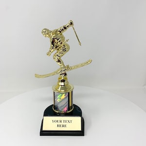 Może przedstawiać: Złoty trofeum przedstawiający narciarza na nartach z czarną podstawą i marmurową, tęczową kolumną. Podstawa ma złotą tabliczkę z tekstem "YOUR TEXT HERE".