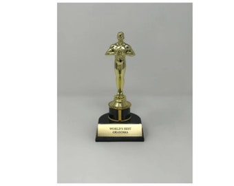 May include: A gold-colored trophy with a figure holding a bouquet, mounted on a black base with a gold plaque. The plaque reads "WORLD'S BEST GRANDMA". The trophy is a decorative award.