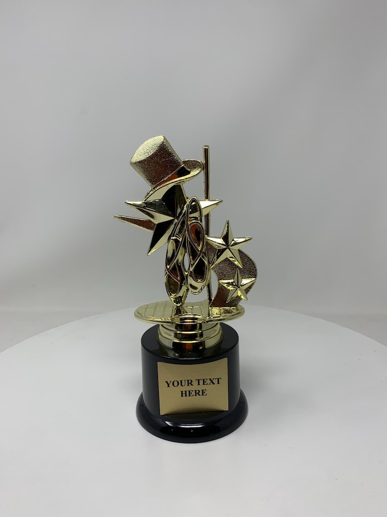 May include: Gold-colored trophy with a top hat, stars, and ballet shoes. The base is black with a gold plate that says "YOUR TEXT HERE".