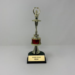 以下が含まれることがあります： 赤い金色の台座が付いた銀色のバレリーナのトロフィー。台座には「YOUR TEXT HERE」という文字が書かれた黒いプレートが付いています。