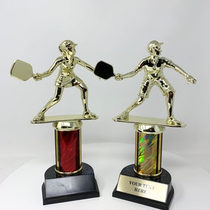 May include: Two gold-colored pickleball player trophies on black bases with red and iridescent columns. The base of the right trophy has the text "YOUR TEXT HERE".