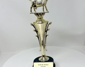 Può includere: Un trofeo dorato con una statuetta di capra in cima a una colonna decorativa. Il trofeo ha una base nera con una placca dorata che recita "YOUR TEXT HERE". Il trofeo è probabilmente per un risultato o un premio.