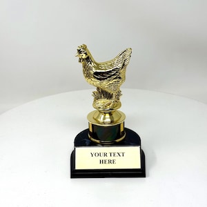 May include: A gold-colored chicken trophy with a black base and a gold plaque that reads "YOUR TEXT HERE". The chicken is perched on a small patch of grass. The trophy is on a white surface.