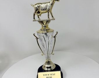 Può includere: Un trofeo dorato con una figura di capra in cima a una base trasparente e dorata. Il trofeo ha una base nera con una placca dorata che recita "YOUR TEXT HERE". Questo trofeo è probabilmente per un premio o un riconoscimento.