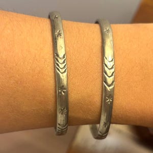 May include: Two silver-toned metal bangles featuring a repeating pattern of engraved star-like shapes and chevron-style arrowheads. The bracelets have a smooth, rounded profile and a matte finish, displaying consistent etched detailing throughout.