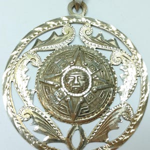 May include: A silver pendant with an Aztec calendar design. The pendant features a central sun face surrounded by intricate patterns and a decorative outer ring. The pendant has a loop for a chain.