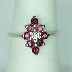 May include: A silver ring featuring a floral design. The ring's centerpiece is a cluster of deep red gemstones surrounding a single, clear gemstone. The ring is set on a white background, highlighting the contrast between the metal and the stones.