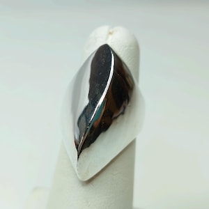 May include: A polished silver ring with a smooth, elongated design. The ring's surface reflects light, creating a bright, shiny appearance. The ring is displayed on a white stand, showcasing its shape and design. A piece of jewelry.