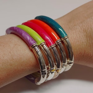 May include: A collection of colorful bangles in shades of purple, green, red, orange, and teal. Each bangle has a silver-colored metal band. The bangles are stacked on a wrist.