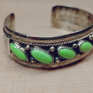 May include: A silver cuff bracelet featuring a row of oval-shaped, bright lime green stones. The metal band is decorated with a twisted rope pattern and small silver beads between each stone, creating a textured, metallic finish throughout.