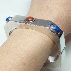 May include: A silver-toned hinged bracelet with three inset gemstones. The gemstones are a deep blue, a reddish-brown, and another deep blue. The bracelet has a geometric design and is worn on the wrist.