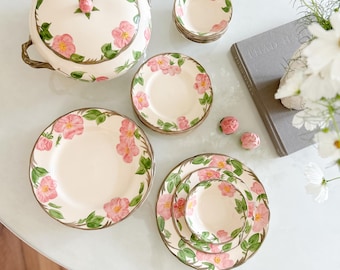 May include: A cream-colored ceramic dinnerware set featuring a floral design with pink roses and green leaves. The set includes a lidded serving dish, plates, and bowls. The rims have a brown trim.