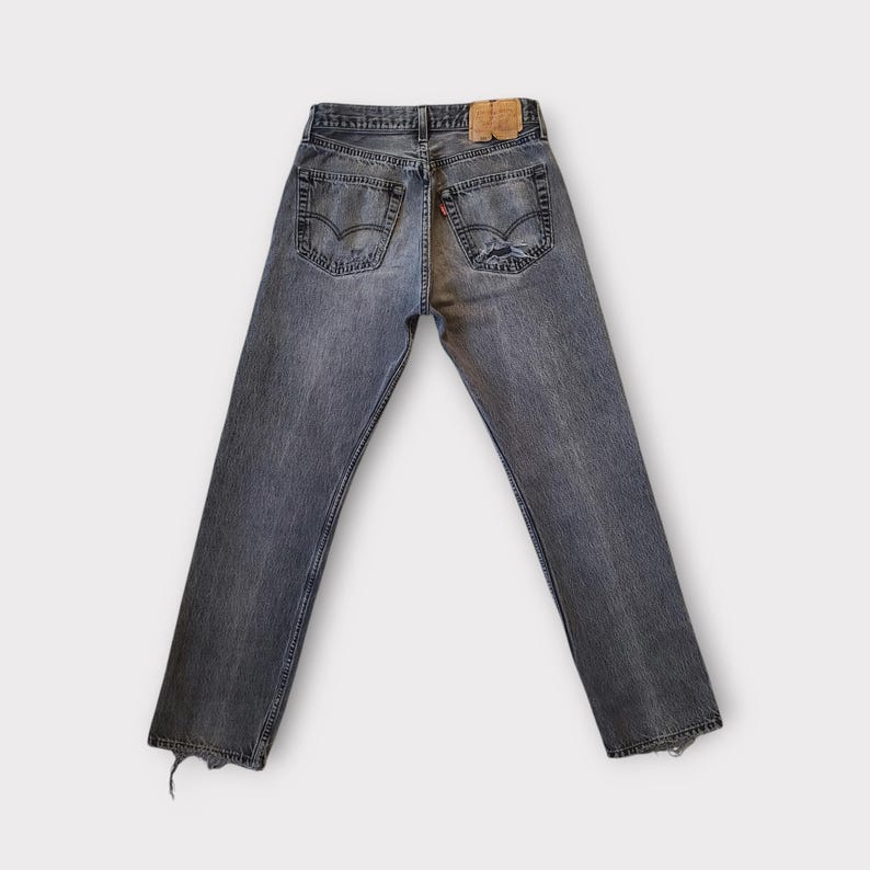 May include: Faded black denim jeans with a distressed look. The jeans have a classic five-pocket design, a leather patch, and a red tag. The bottom hems are frayed. The jeans are displayed on a white background.