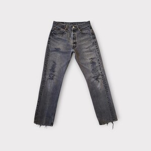 May include: Faded black denim jeans with distressed details, including rips and frayed edges. The jeans have a button fly and classic five-pocket styling. The denim appears worn and softened.