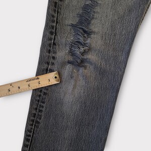 May include: Close-up of distressed denim jeans with a large ripped section. A wooden ruler with inch markings is placed along the edge of the jeans. The jeans are a faded blue color.