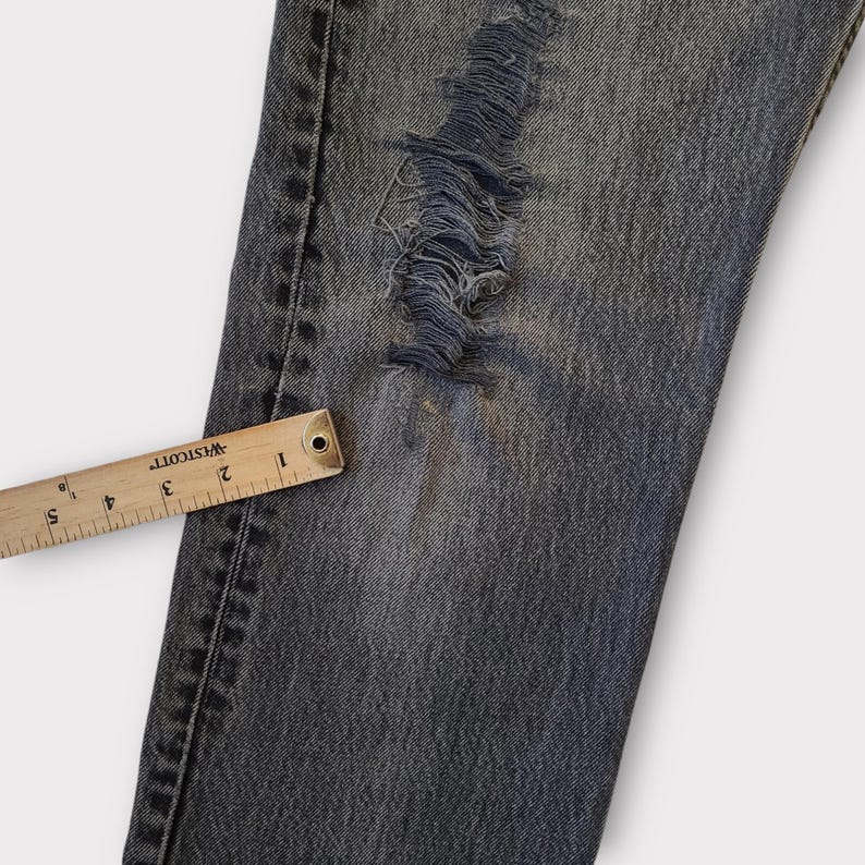 May include: Close-up of distressed denim jeans with a large ripped section. A wooden ruler with inch markings is placed along the edge of the jeans. The jeans are a faded blue color.