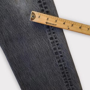May include: Close-up of a section of dark gray denim jeans with a wooden ruler measuring the fabric. The ruler shows measurements in inches. The jeans have visible stitching along the seam.