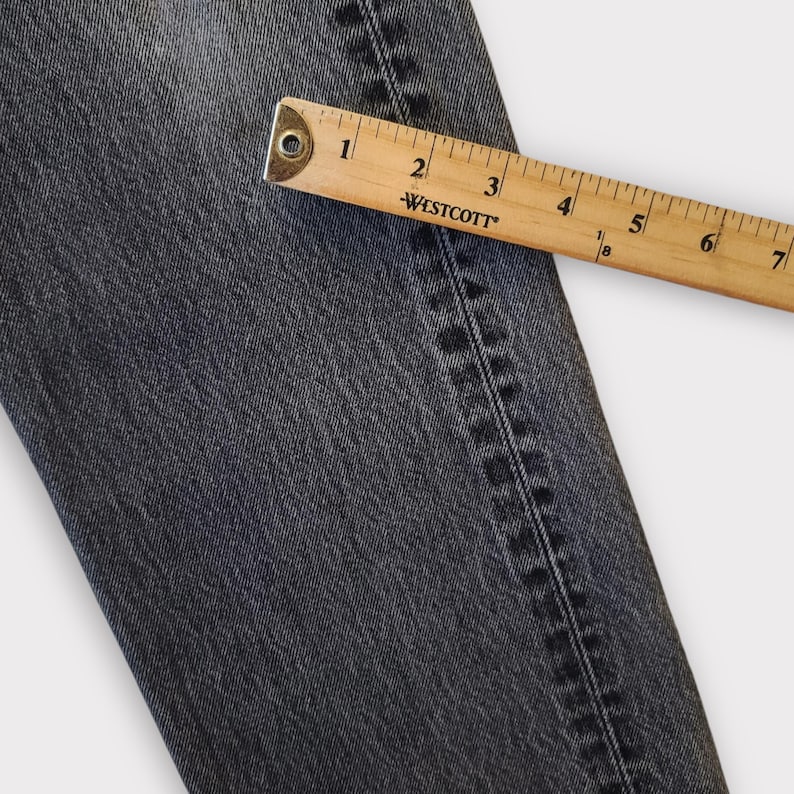 May include: Close-up of a section of dark gray denim jeans with a wooden ruler measuring the fabric. The ruler shows measurements in inches. The jeans have visible stitching along the seam.
