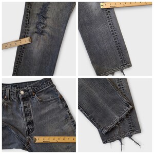 May include: Close-up shots of distressed, faded black denim jeans. The jeans feature ripped details on the leg and a raw, frayed hem. A ruler is placed on the jeans to show measurements. The jeans have a button closure and classic five-pocket design.