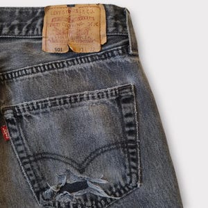 May include: Close-up of a pair of faded black denim jeans. The jeans feature a distressed pocket with a tear, a red Levi's tag, and a brown leather patch with text. The jeans are size 31x32.
