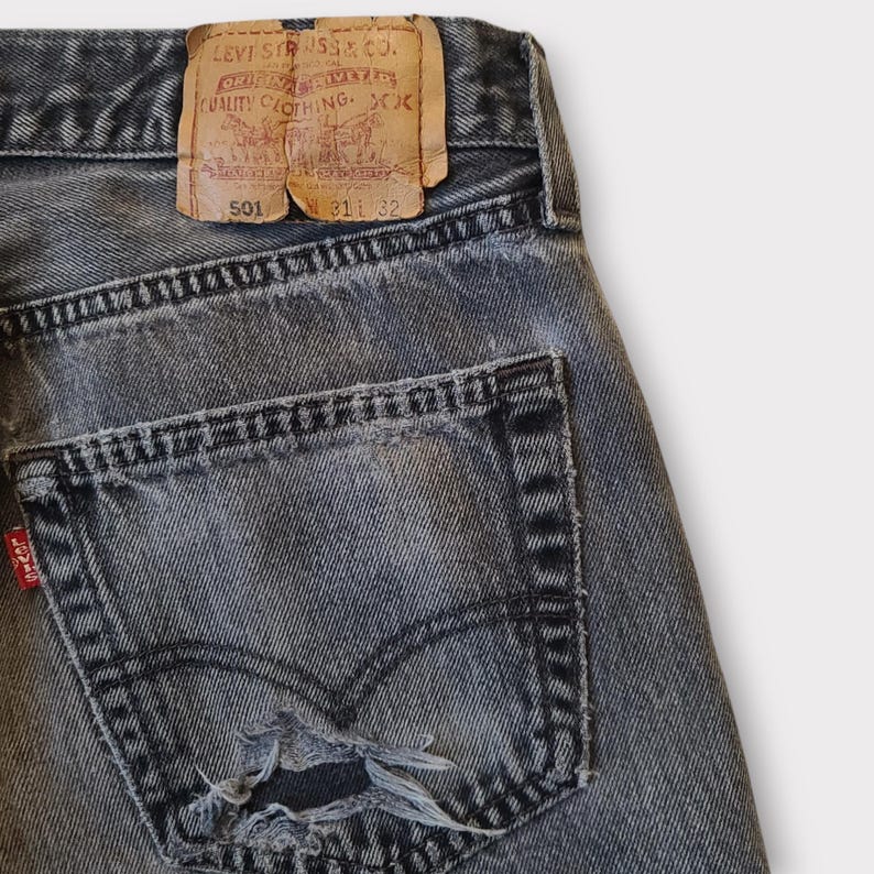 May include: Close-up of a pair of faded black denim jeans. The jeans feature a distressed pocket with a tear, a red Levi's tag, and a brown leather patch with text. The jeans are size 31x32.